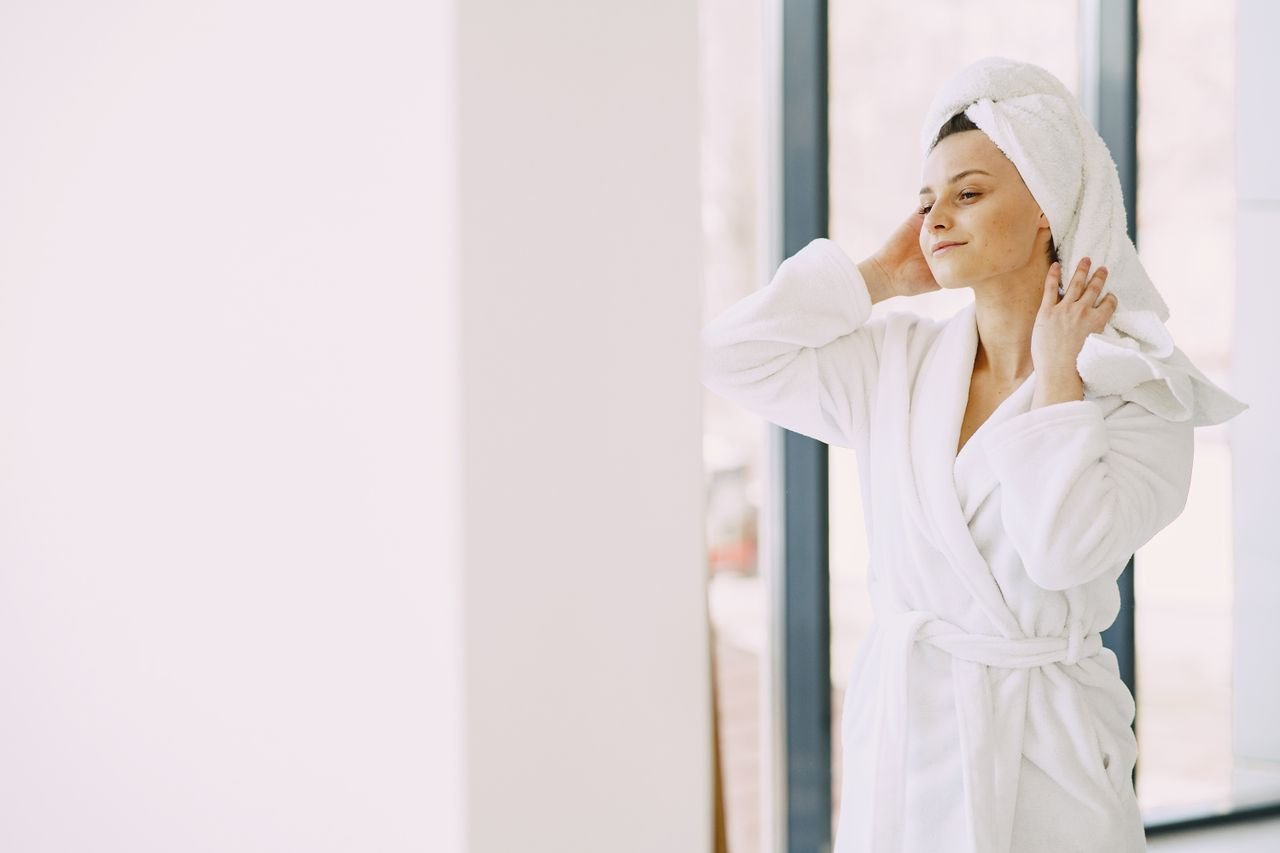 Pensive female in white bathrobe and towel on head smiling and standing near glass window in living room while looking away