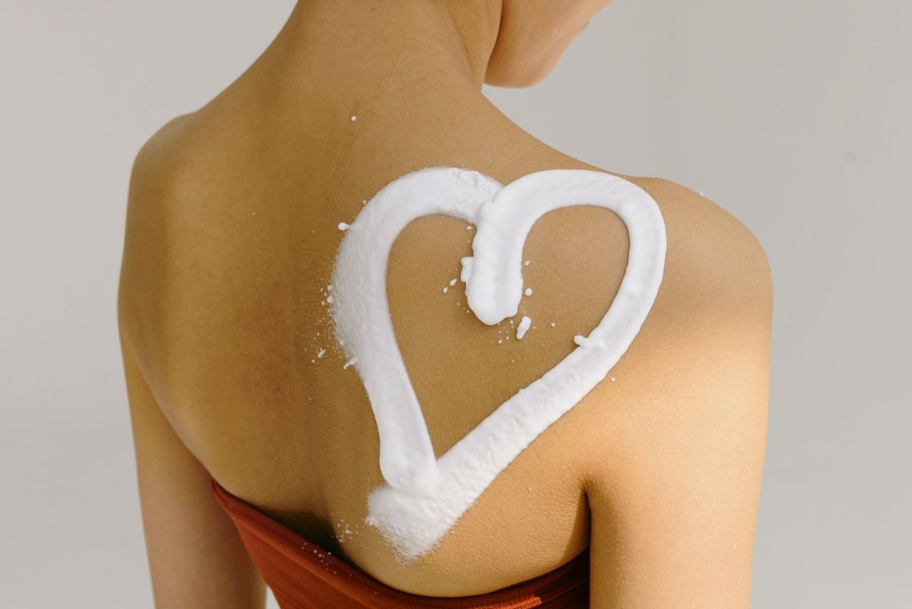 Rear view of a woman's shoulder with a heart shape drawn in cream, symbolizing love and skincare.